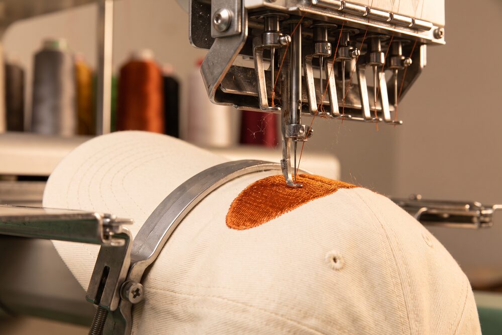 A machine needle stitching dense orange thread into a cap panel
