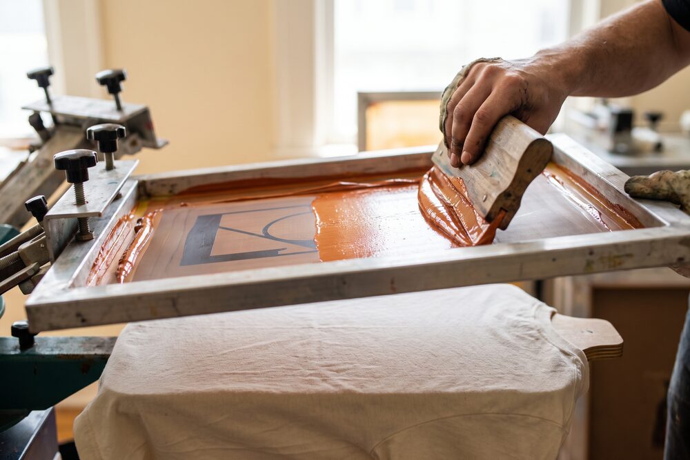 A squeegee pulling orange ink through a mesh screen onto a cotton tee
